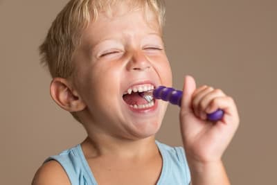 A blonde boy smiles and brushes his teeth with a purple children's toothbrush